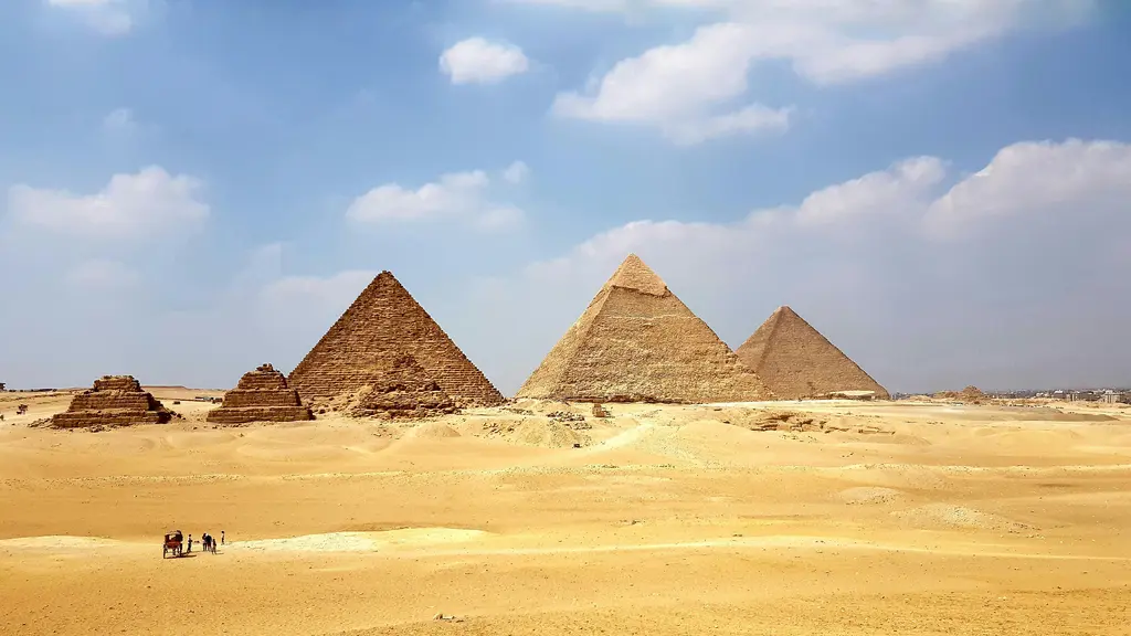 Mısır'da Giza Piramitleri; çölün üzerinde yükselen üç büyük piramit ile önlerindeki kraliçe piramitleri, altın rengi kumda küçük bir at arabası ve mavi gökyüzünde dağınık bulutlar.