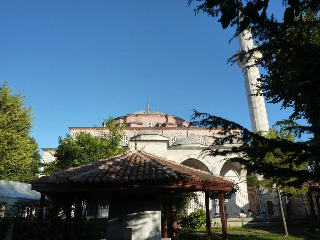Hüseyin Ağa Medresesi'nin bahçesinde kırmızı kiremitli ahşap kameriye; arkada caminin kubbesi ile revağı, sağda sedirin arkasından yükselen minare, lacivert gök ve yeşil çim.