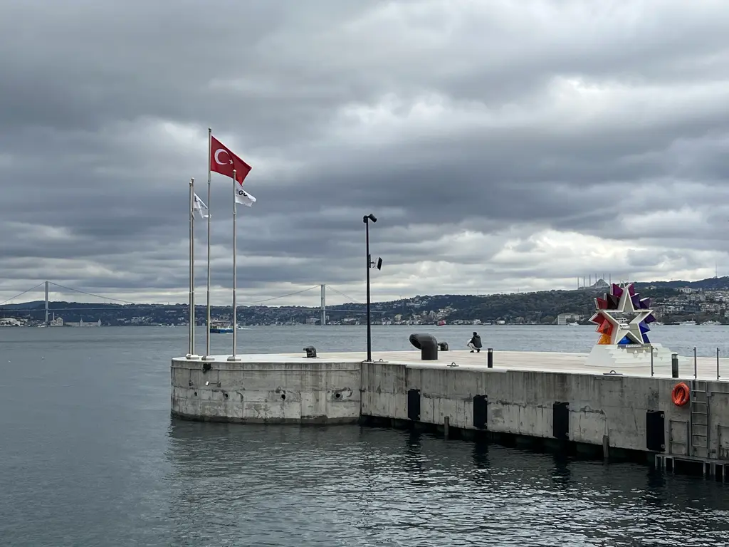 Galataport'ta iskele; bayrak direklerinde Türk bayrakları, iskelenin ucunda heykel, arkada Boğaz ve uzakta Boğaziçi Köprüsü.
