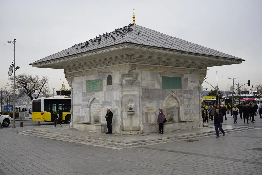 Güneyden III. Ahmed Çeşmesi; çatıda güvercinler, mermer gövde, köşe muslukları, arkada otobüsler ve deniz.