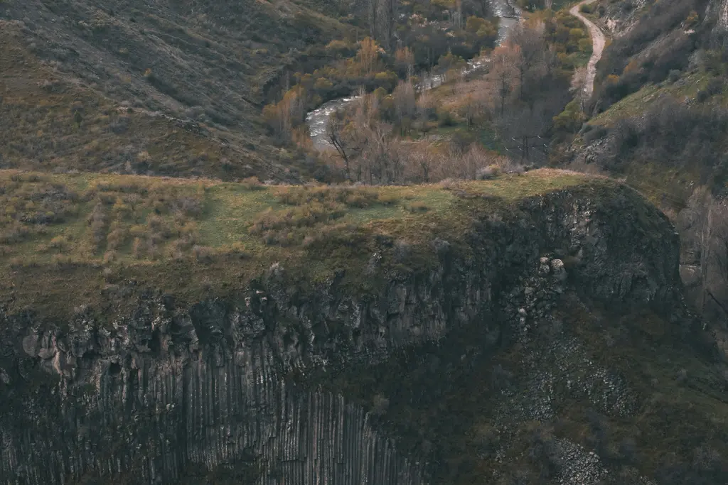 Garni Vadisi'nde kanyon duvarını oluşturan düzgün altıgen bazalt sütunlar; üstte yeşil çayırla kaplı plato, aşağıda ağaçların arasından kıvrılan nehir ve toprak yol.