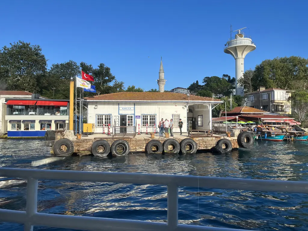Vapurdan Kanlıca İskelesi; lastik kaplı rıhtım, iskele binası, arkada minare, beyaz radar kulesi ve mavi gökyüzü.