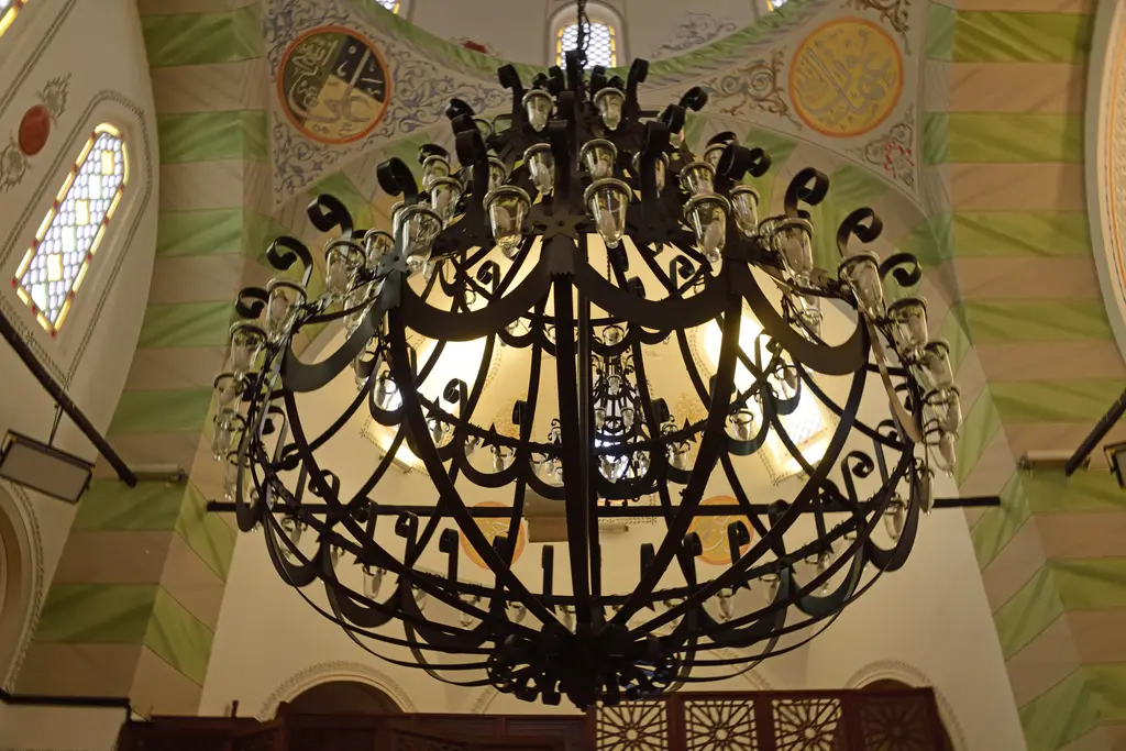Koca Mustafa Paşa Camii'nde dev dövme demir avizeye alttan bakış; halka gövde, yukarı bakan ampuller, kıvrık demir kollar, arkada yeşil bantlı ve altın madalyonlu kalem işi tonoz.