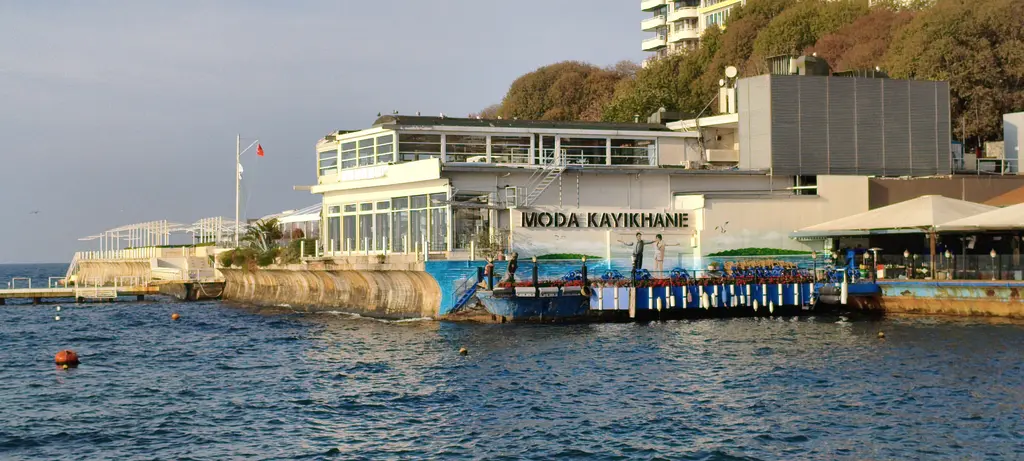 Moda Kayıkhanesi; denize kurulu beyaz bina, iskeleye bağlı tekneler, yamaçta ağaçlar ve apartmanlar.