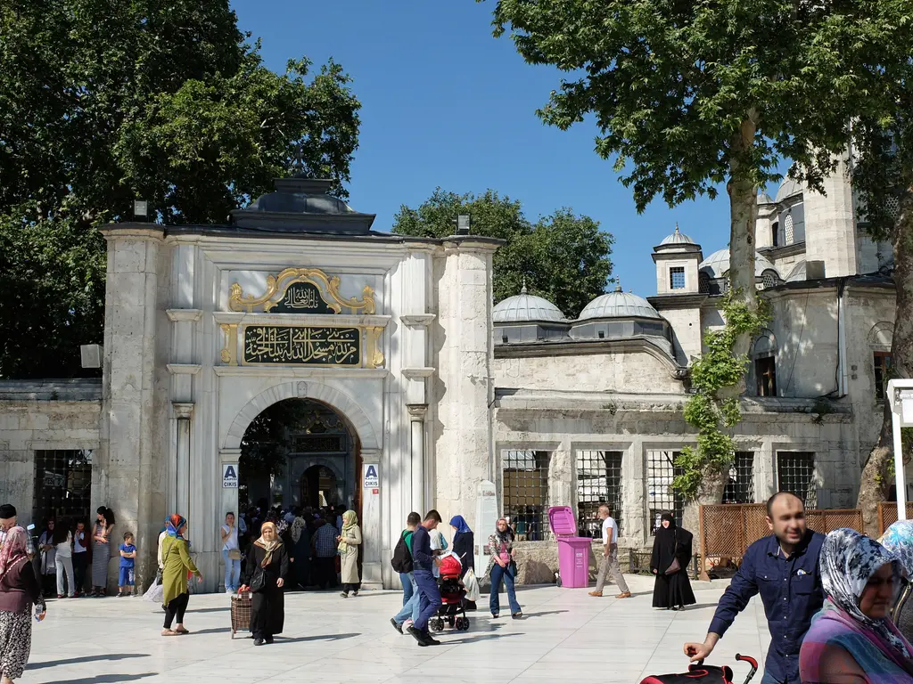 Eyüp Sultan Camii'nin dış avlu kapısı; kitabeli mermer taç kapı, yaldızlı süslemeler ve önünde ziyaretçiler.