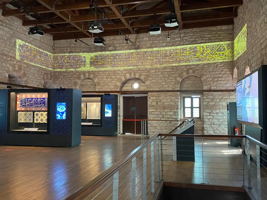 Tekfur Sarayı'nın üst kat sergi salonu; ahşap tavan, duvar boyunca çini desenli friz, dijital ekranlar ve önde vitrinli sergi üniteleri.