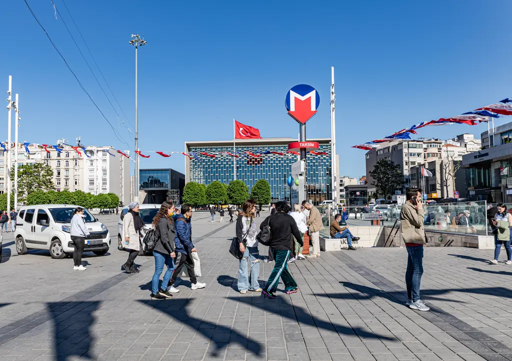 Taksim Meydanı'nda metro girişini gösteren kırmızı M tabelası; önde yürüyen insanlar, arkada Türk bayraklı Atatürk Kültür Merkezi.