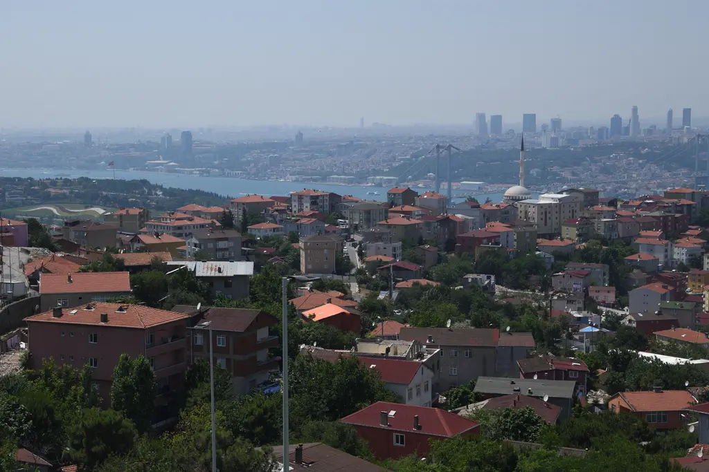 Çamlıca'dan manzara; ön planda yeşil yamaç ve çatılar, uzakta Boğaz, köprü ve Avrupa yakasının kuleleri.