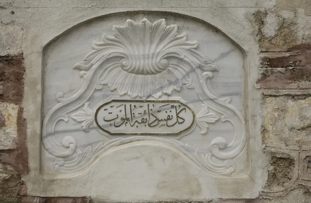 Atik Valide Camii'nde mermer kitabe; kıvrık yapraklı barok çerçeve içinde oval madalyonda Arapça yazı.