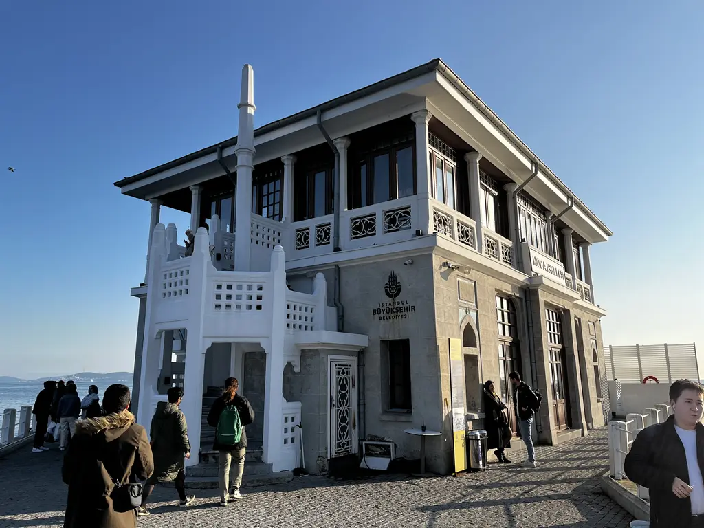 Moda İskelesi'nin kara cephesi; gri taş duvar, beyaz dış merdiven, üst katta sütunlu galeri ve önde yürüyen insanlar.