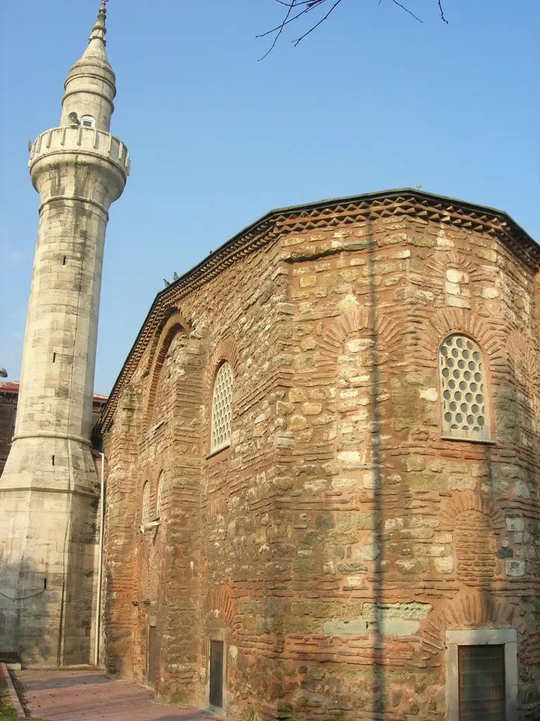 Atik Mustafa Paşa Camii'nin taş minaresi ile tuğla gövdesi mavi gökyüzü altında; tek şerefeli ince minare, yanında çok yüzlü duvar ve kemerli şebekeli pencereler.