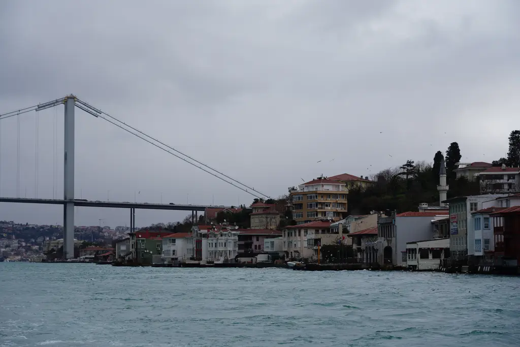 Kuzguncuk'ta Boğaz'dan kıyı; suya inen beyaz ve renkli yalılar, arkada yamaçtaki evler ve solda asma köprü.