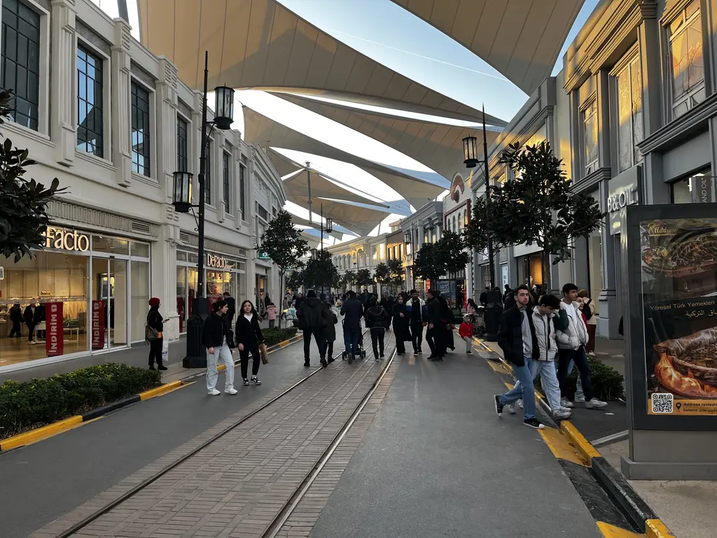 Vialand alışveriş caddesinde üstü beyaz gölgelik yelkenleriyle örtülü yol; ortada tramvay rayları, iki yanda mağazalar ve kalabalık.