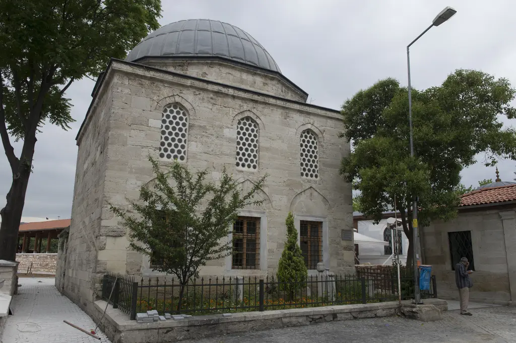 Avlunun arkasındaki kubbeli kâgir yapı; taş duvarlar, küçük pencereler, çevresinde ağaçlar ve önünde demir korkuluk.