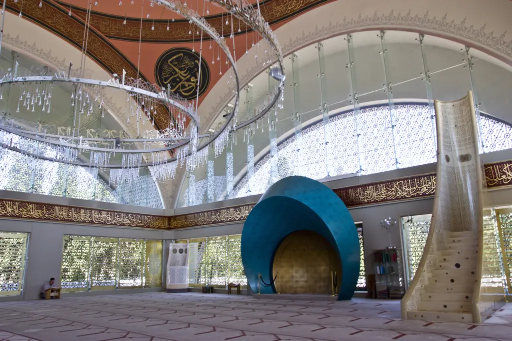 Şakirin Camii'nin harimi; turkuaz mihrap, beyaz akrilik minber, kubbeden sarkan halkalı avize ve cam cepheler.