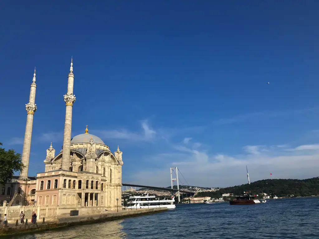 Ortaköy Camii; koyu mavi gökyüzü altında açık renkli taş cephe, iki ince minare, sağda köprü ve suda bir yolcu teknesi.
