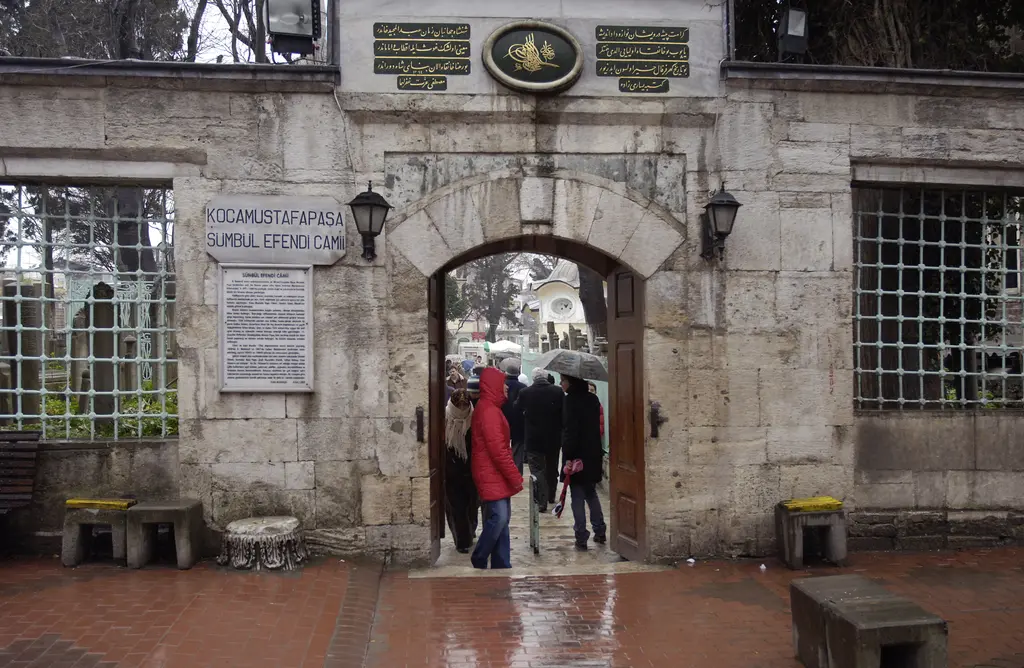 Koca Mustafa Paşa Camii'nin kitabeli kapısı yağmurlu bir günde; kemerin üstünde üç yeşil zeminli Osmanlıca levha ile altın tuğra, iki yanda şebekeli pencereler, ıslak kırmızı tuğla döşeme.