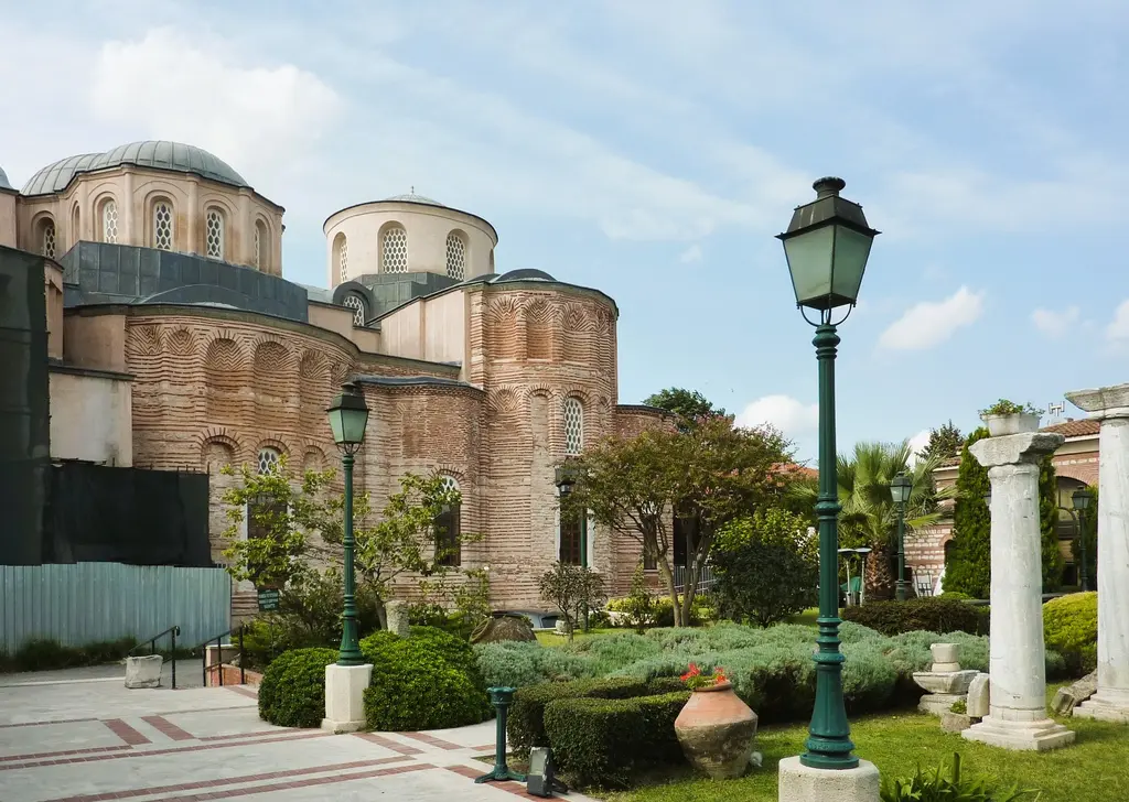 Zeyrek Camii'nin bahçesi; taş döşeli yol, yeşil sokak lambaları, çimende duran antik sütun parçaları ve arkada birbirine bitişik kubbelerle tuğla taş örgülü cephe.
