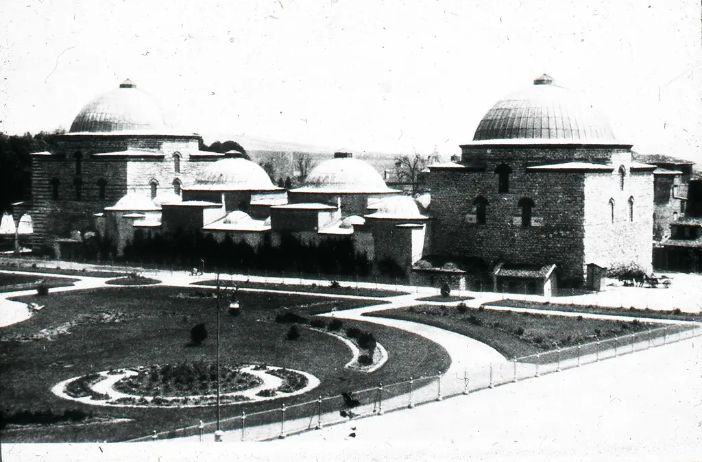 Eski bir fotoğrafta hamamın park cephesi; önde geometrik yollarla bölünmüş düzenli bahçe, arkada kubbeler ve alçak çatılar.