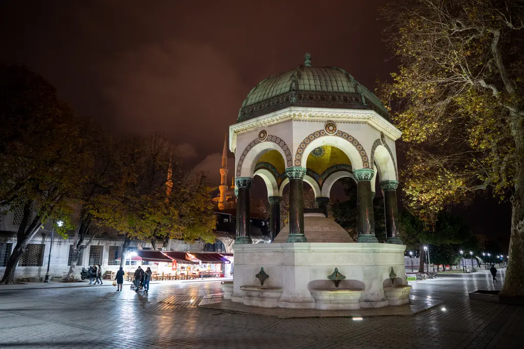 Akşam Alman Çeşmesi; aydınlatılmış kemerler, koyu kubbe, arkada ışıklı ağaçlar, ıslak taş zeminde yansımalar ve uzakta dükkân ışıkları.