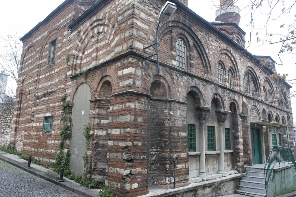 Vefa Kilise Camii'ne kuzeybatı köşesinden parke sokaktan bakış; kararmış almaşık duvar, sağır kemerler, sarmaşık, sağda mermer sütunlu revak ve yeşil pencere doğramaları.