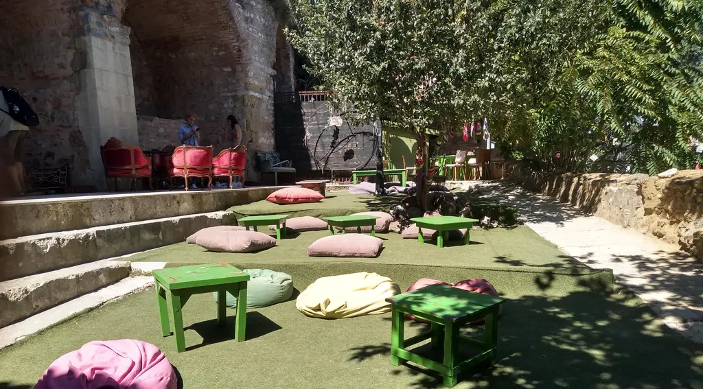 Balat'ta eski taş duvar dibinde bahçe kafe; çimlerde minderler, yeşil masalar ve ağaç gölgesi.