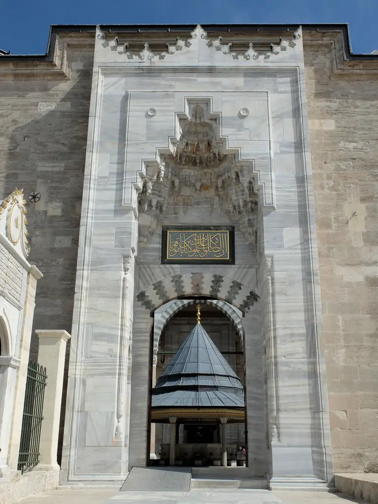 Fatih Camii'nde avlunun mermer taç kapısı; damarlı gri mermer çerçeve, altın hatlı siyah kitabe panosu, üstte mukarnas kavsara ve açıklıktan görünen şadırvan.
