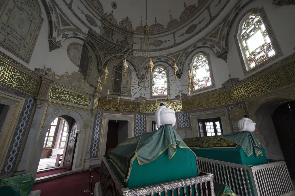 III. Mustafa Türbesi'nin içi; yeşil örtülü sandukalar, uçlarında sarıklar, çevresinde pirinç şebeke ve duvarlarda çini panolar.