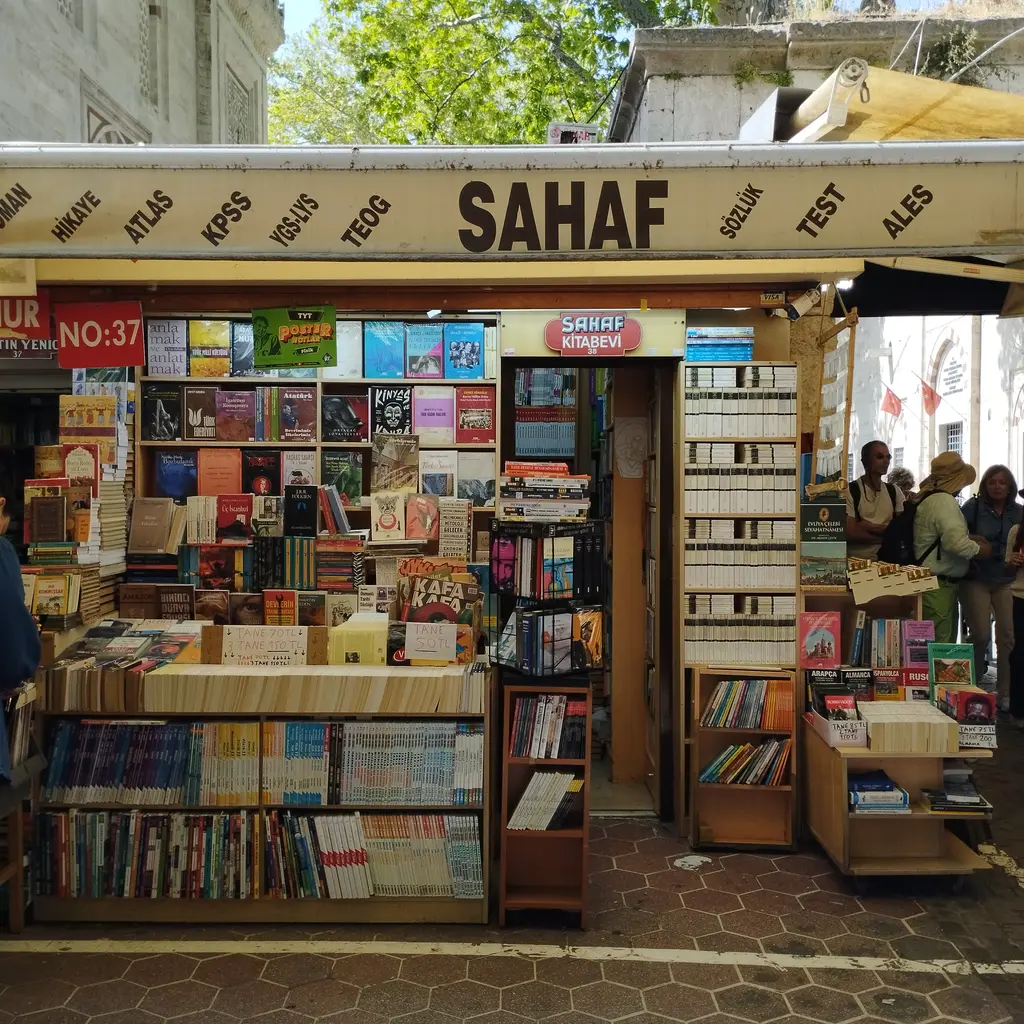 Sahaflar Çarşısı'nda bir sahaf dükkânının önü; sarı zeminde siyah "Sahaf" tabelası, yanında kitap türlerini sayan sütunlar, ahşap raflar ve parkeye kadar taşmış kitaplar.