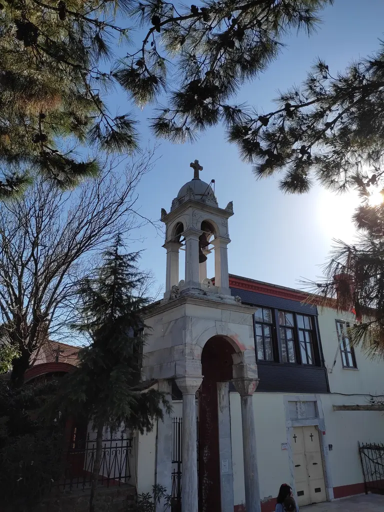 Aya Yorgi'nin çan kulesi; çam dallarının arasından beliren kemerli kule ve ahşap çıkmalı manastır binası.