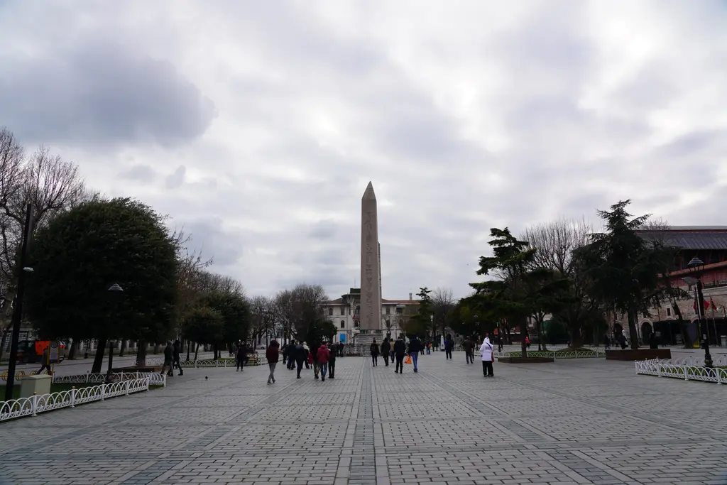 Sultanahmet Meydanı'nın geniş taş döşeli yaya alanı; ortada Dikilitaş, iki yanda ağaçlar, yürüyen insanlar ve bulutlu gökyüzü.