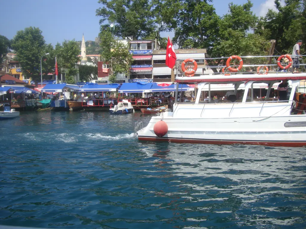 Anadolu Kavağı'nda limana giren beyaz tur teknesi; can simidi asılı küpeşte, arkada mavi tenteli lokantalar ve minare.