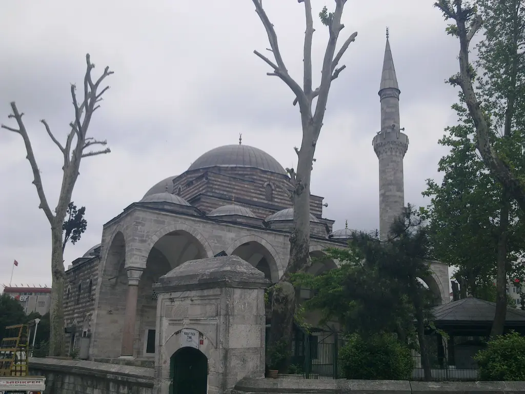 Murat Paşa Camii'ne avlu kapısından bakış; taş kapı, kubbeli cami ve tabhane kubbeleri, arkada minare ve kuru dallı ağaçlar.