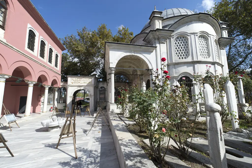Şah Sultan Külliyesi'nin avlusu; pembe mektep binası, güller, mezar taşları ve arka planda kubbeli türbe.