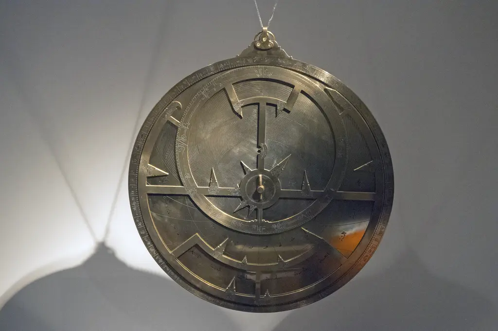Büyük bir usturlap kopyası; gri fonda asılı yuvarlak metal disk, üzerinde oyma tutamaç, kesişen çizgiler ve kenar işaretleri.