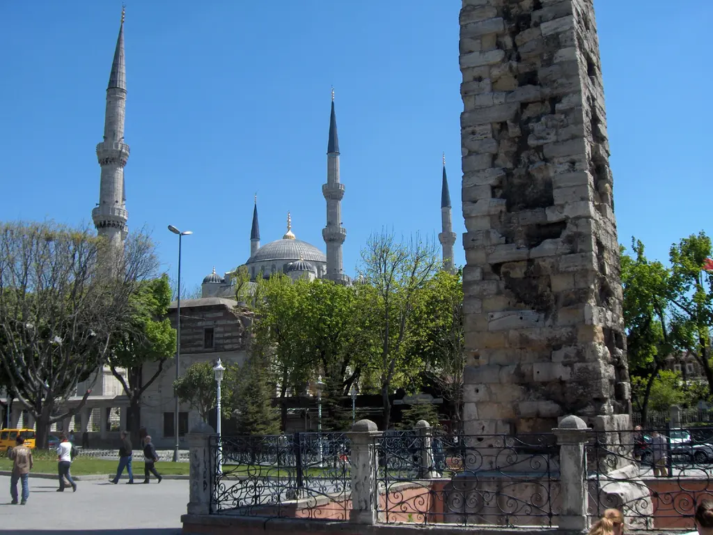 Örme Sütun'un yıpranmış taş gövdesi, arkada Sultanahmet Camii'nin kubbeleri ve minareleri, önde parmaklık ve yeşil ağaçlar.