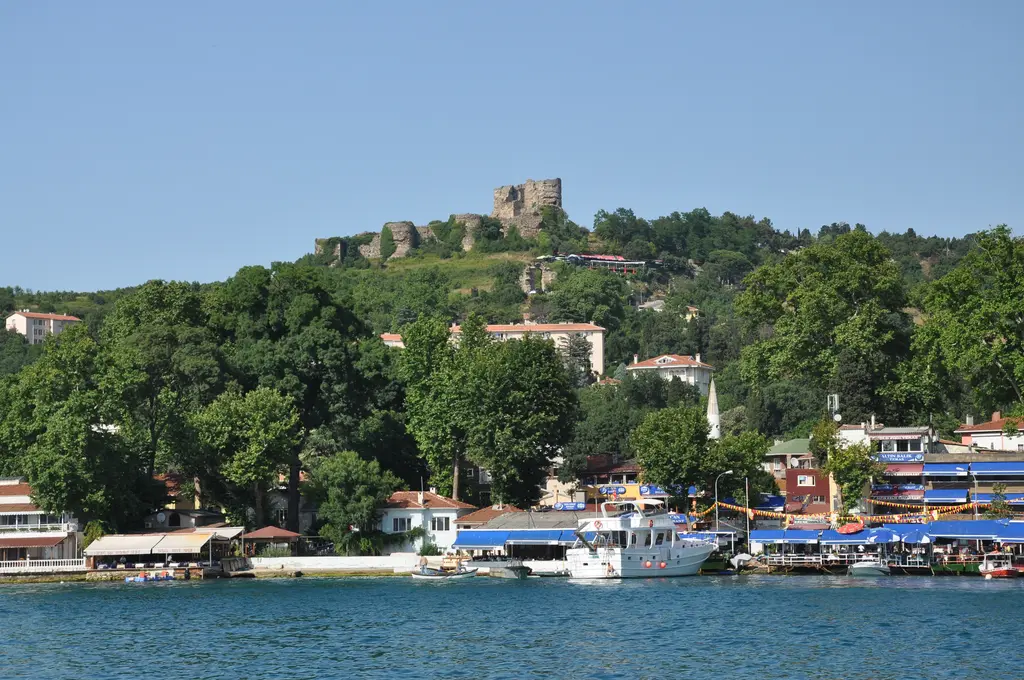 Anadolu Kavağı'nda Boğaz'dan köy; kıyıda mavi tenteli lokantalar ve tekneler, ağaçlı yamaçta evler ve tepede Yoros Kalesi.