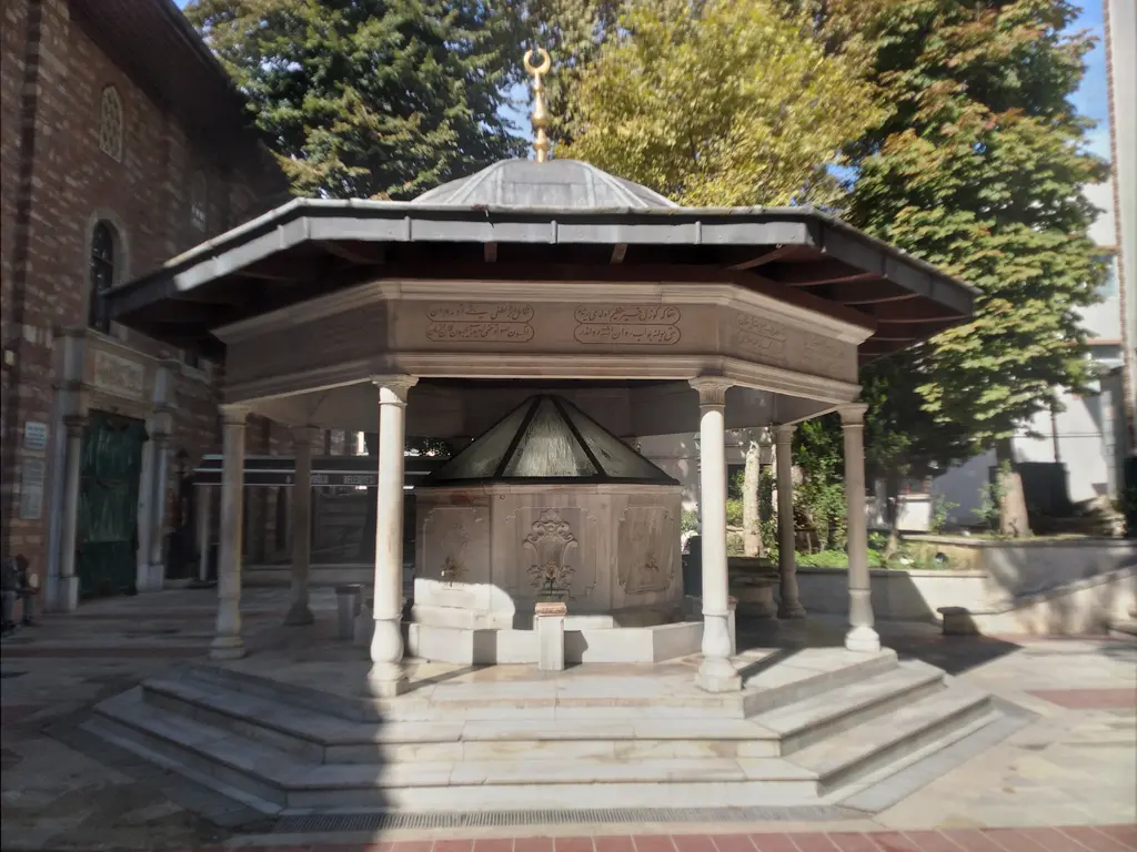 Arap Camii'nin şadırvanı; ince sütunlara oturan geniş saçaklı kubbeli çatı, mermer hazne, basamaklar ve arkada ağaçlar.