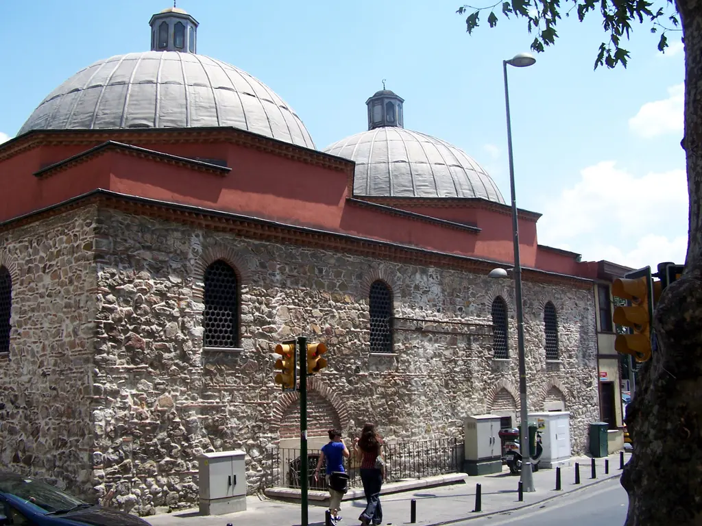 Hüsrev Kethüda Hamamı'na köşeden bakış; iki büyük kubbe, taş ve tuğla örgü duvarlar, kırmızı saçak ve mavi gökyüzü.