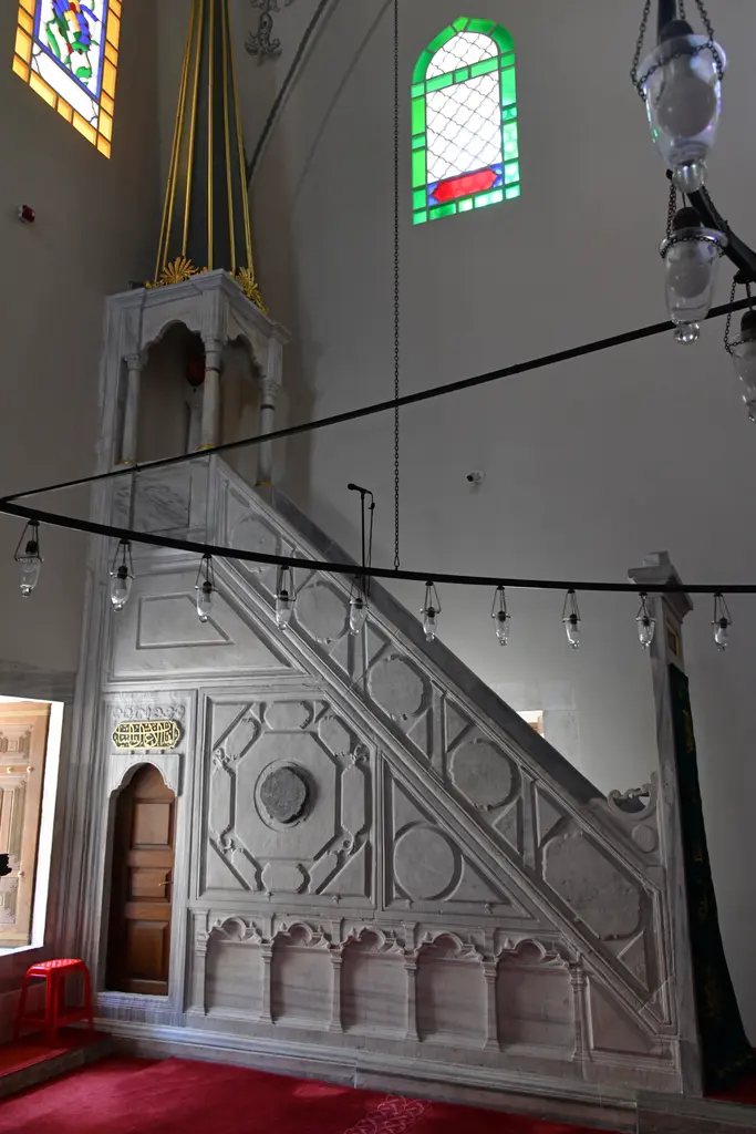 Mahmut Paşa Camii'nin minberi; geometrik kabartmalı mermer korkuluk, sivri külah, kemerli kapı, arkada renkli camlı pencere ve kandiller.