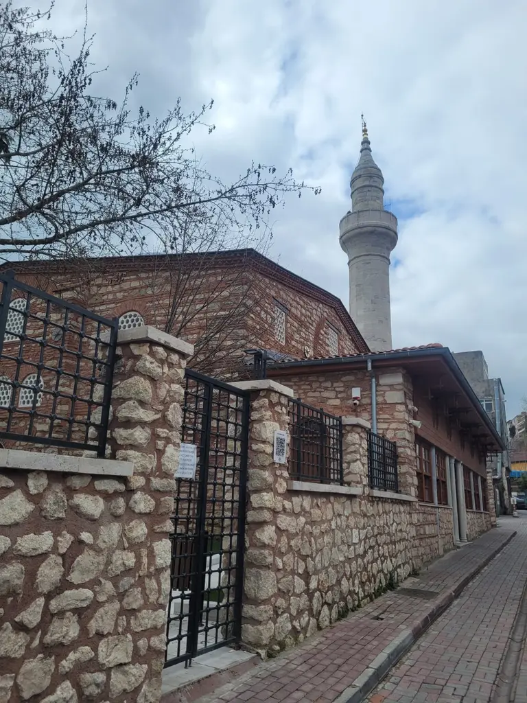 Atik Mustafa Paşa Camii'nin Çember Sokak cephesi; moloz taş duvar üstünde siyah demir parmaklık, açık demir kapı, arkada tuğla gövde ile minare ve kırmızı parke kaldırım.