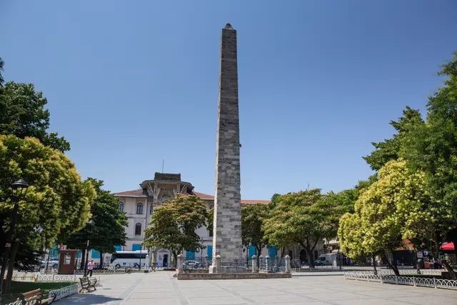 Sultanahmet Meydanı'nda Örme Sütun; kaba taş örgülü uzun gövde, çevresinde yeşil ağaçlar, arkada binalar ve açık mavi gökyüzü.