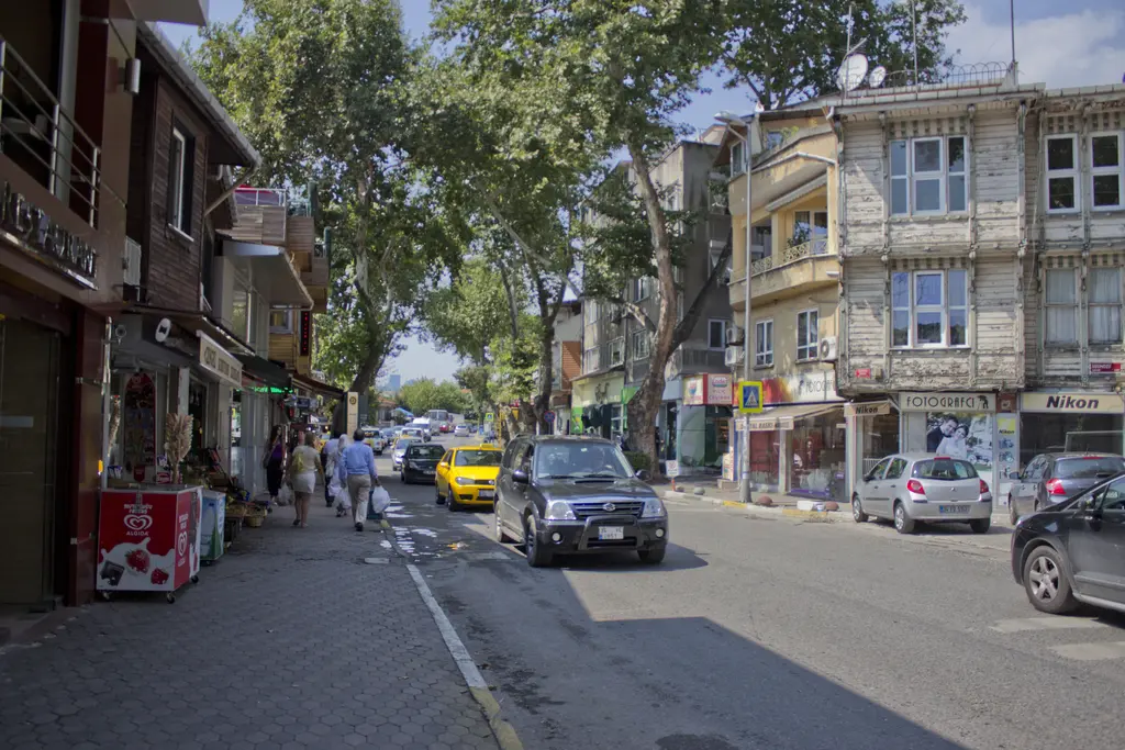 Çengelköy'ün ana caddesi; çınar ağaçları altında dükkânlar, ahşap kaplı eski ev, kaldırımda yürüyenler ve arabalar.
