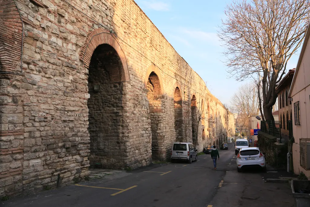 Bozdoğan Kemeri'nin mahalle içinden geçtiği dar sokak; solda kalın kemer ayağı ve tuğla kuşaklı büyük açıklık, sokakta beyaz kamyonet, sağda pastel renkli apartmanlar.