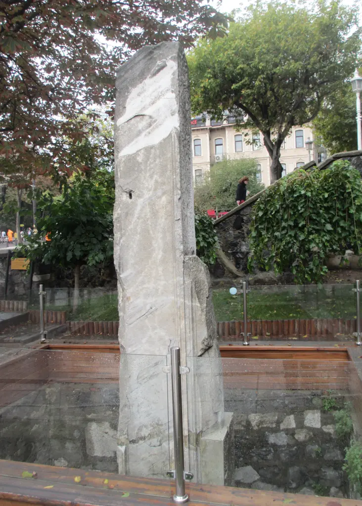 Milyon Taşı; ahşap kenarlı, cam korkulukla çevrili çukurda duran mermer ayak, arkada yeşil ağaçlar, merdiven ve eski binalar.