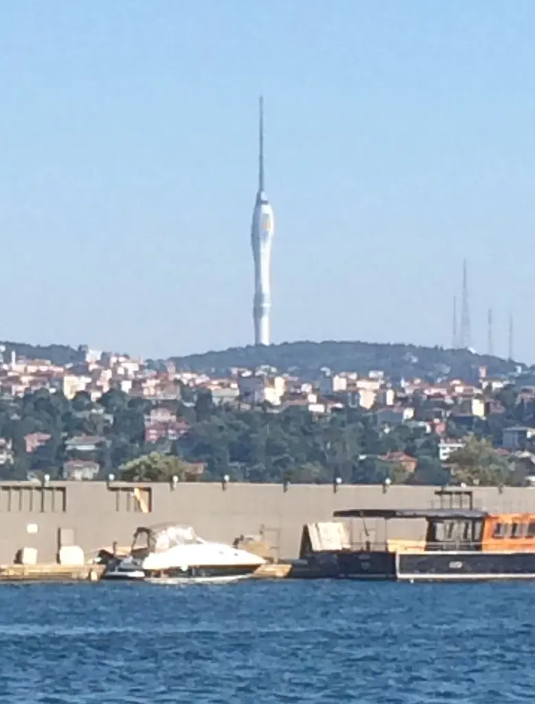 Denizden Çamlıca Kulesi; önde Boğaz kıyısındaki yapılar ve tekneler, yamaçta yerleşim ve tepede kule.