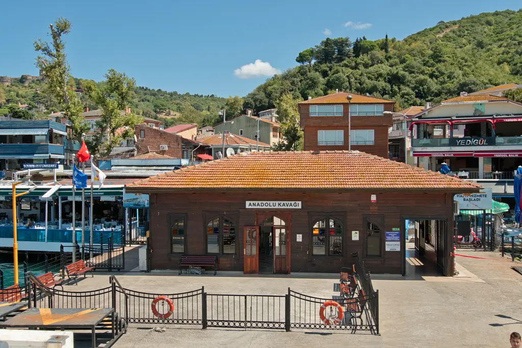 Anadolu Kavağı iskelesi; kiremit çatılı ahşap iskele binası, demir korkuluklu meydan ve arkada lokantalar ile yeşil tepe.