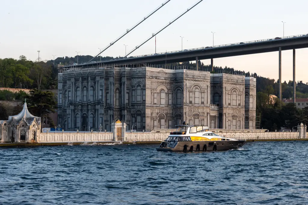 Boğaz'dan Beylerbeyi Sarayı; deniz cephesi, solda çadır çatılı deniz köşkü, üstünden geçen köprü ve önünde bir tekne.