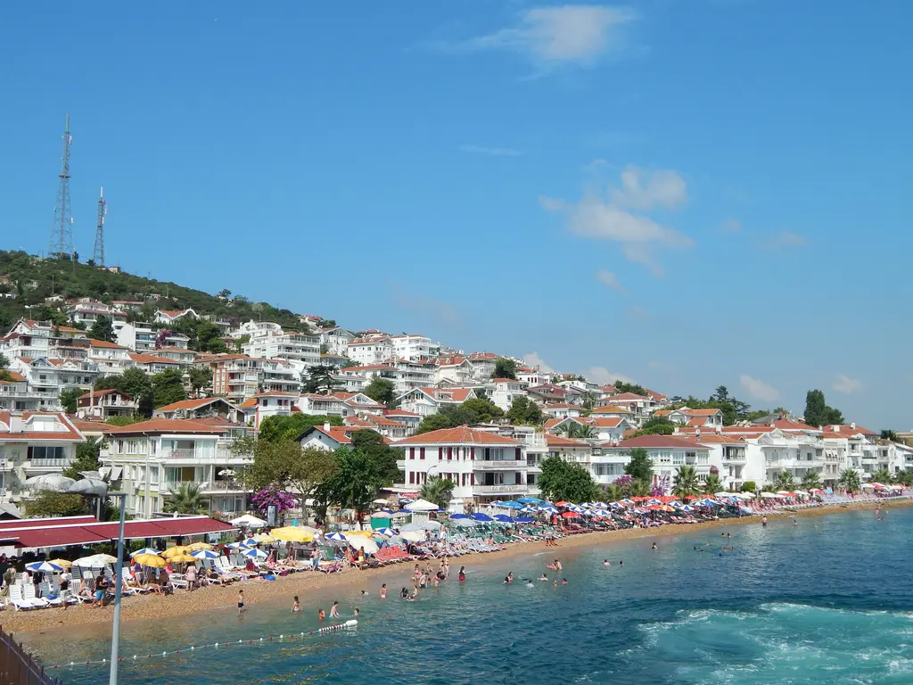 Kınalıada kıyısı; tepeye tırmanan beyaz evler, kıyıda dolu bir halk plajı ve iskeleye yanaşan tekneler.