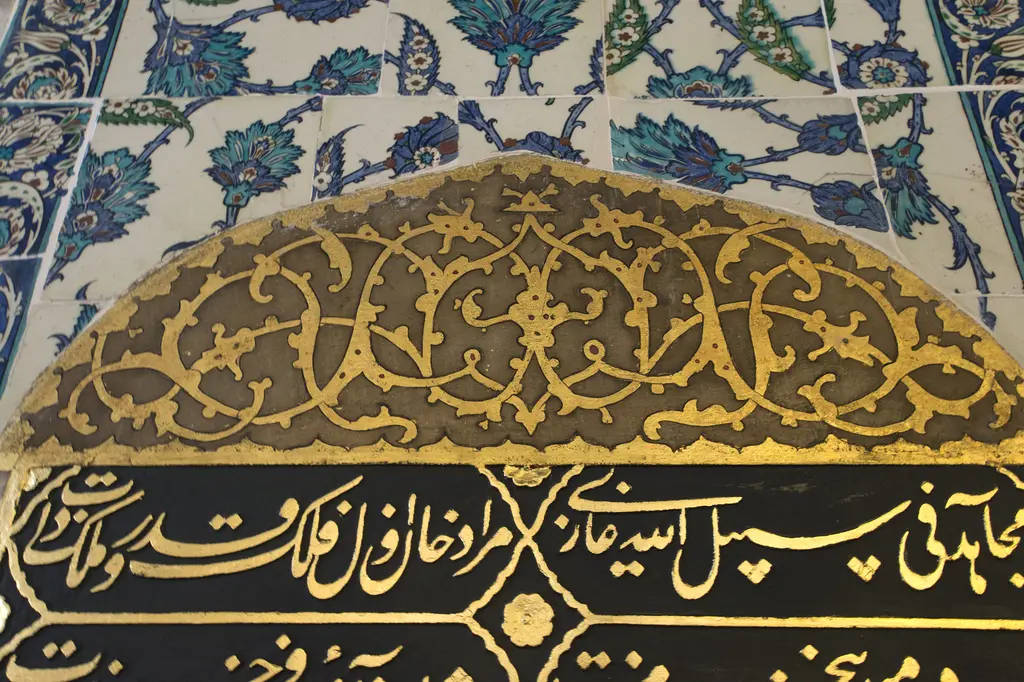 Sultan Ahmed Türbesi'nde yazılı pano; çini kaplı duvarın altında altın yaldızlı kıvrık dal süslemeli kemer ve siyah zemin üzerine altın yazı.